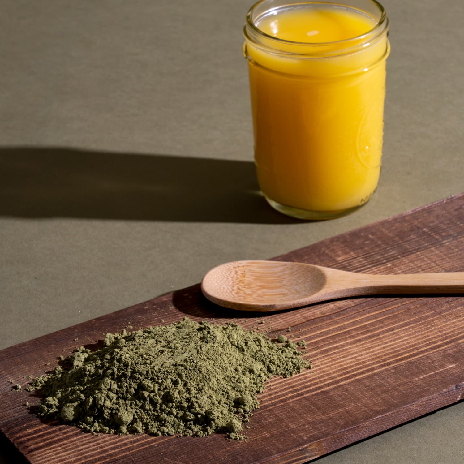 A wooden cutting board with a spoon and brown powder next to a glass of orange juice